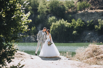 A young groom in a gray tailcoat with a black bow tie and a beautiful, curly-haired bride in a white long dress embrace and kiss, holding hands near the river and rocks. Wedding portrait of newlyweds.