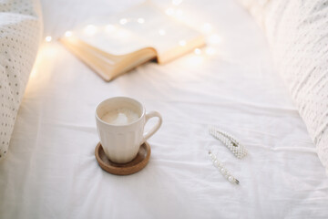 Coffee cup with milk foam and a book on a white bedsheet. 