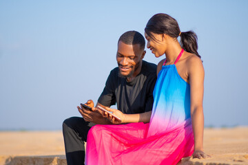 young african man and woman viewing content on a phone together outdoor on a beach, sitting on the...