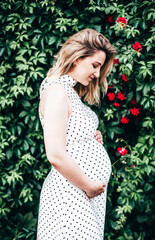 a pregnant fair-skinned woman of European appearance in a light dress against a background of greenery and flowers