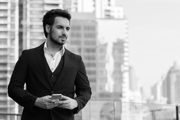 Young handsome Indian businessman holding mobile phone while thinking at rooftop against view of the city