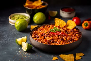 Poster Mexican hot chili con carne in a bowl with tortilla chips on dark background © petrrgoskov