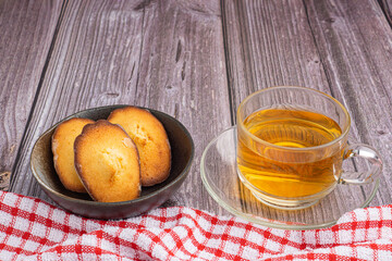 Madeleine homemade traditional French small cookies on black a dish with a cloth and a teacup placed on a wooden background. Lemon glaze madeleines. Seashell sweet cakes