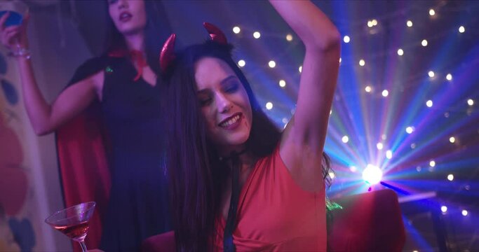 Medium Shot, Handheld &ndash; A beautiful sexy Caucasian lady dressed in red she devil dancing and enjoying a Halloween party with friend in a dark room with decorative lights. 