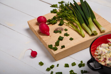 Radish, green onions, parsley, eggs salad in red bowl. Some fresh vegetables and greenery on kitchen chopping board on white wooden background. Diet and nutrition concept.