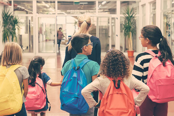 Group of children with female teacher walking in school corridor. Back view. Education or back to school concept