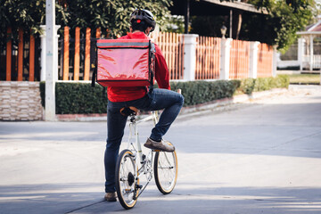 A courier in red uniform with delivery box on back riding bicycle and looking on cellphone to check addres to deliver food to customer. Courier on bicycle delivering food in city.