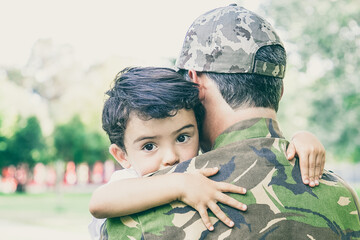 Back view of middle-aged father holding and embracing his son. Lovely little boy hugging dad in army uniform and looking away. Greenery on background. Childhood, weekend and military parent concept