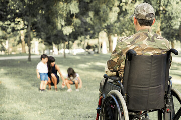 Lonely disabled retired military man in wheelchair looking at his wife and little children playing together in park. Back view. Veteran of war or disability concept