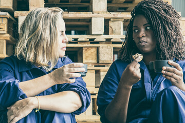 Diverse female factory workers drinking coffee, eating cookies, sitting at wooden pallets and...