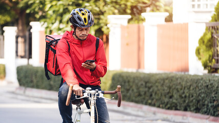 Naklejka premium A courier in red uniform with delivery box on back riding bicycle and looking on cellphone to check addres to deliver food to customer. Courier on bicycle delivering food in city.