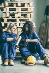 Fototapeta na wymiar Diverse female factory workers sitting on wooden pallet on plant floor, drinking coffee, pouring beverage from thermos to mug. Front view. Industrial occupation or coffee break concept