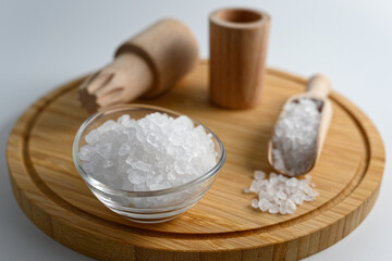 Little wooden scoop and glass bowl full of sea salt on wooden cutting board on white background