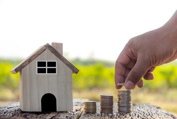 An Asian man's hand placed coins on a pile next to a wooden house on a wooden table. Ideas of...