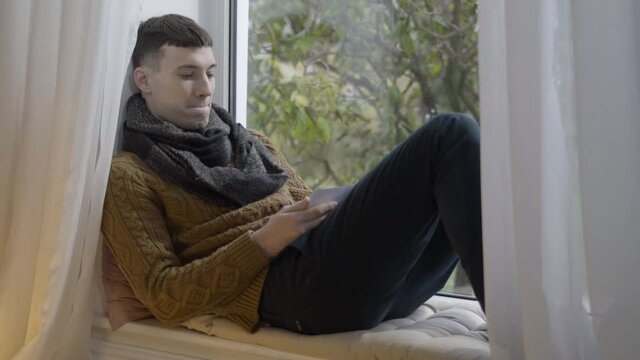 Portrait of lonely depressed man looking at photo sitting on windowsill at home. Frustrated desperate young Caucasian guy putting picture on chest and looking out the window thinking.