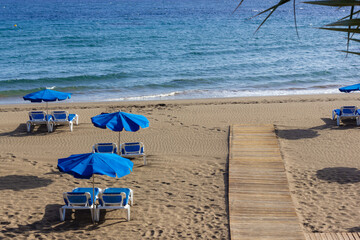 Wooden pathway leading to seashore with blue hammocks and umbrellas on empty beach in Puerto del Carmen, Lanzarote. Tourism abscence, Covid-19 lockdown, economy decline concepts