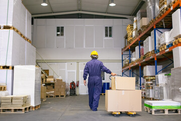 Senior logistic worker in hardhat and uniform walking in warehouse, wheeling palette jack. Back view, full length. Labor and logistics concept