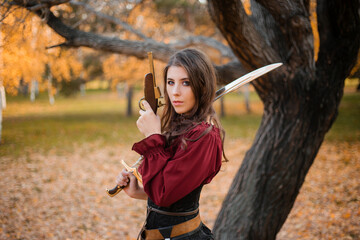 A beautiful girl in a shirt, corset and tight trousers with a saber and a pistol in her hands against the background of an autumn park in the evening. Adventurer woman concept.