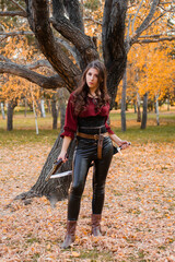 A beautiful girl in a shirt, corset and tight trousers with a saber and a pistol in her hands against the background of an autumn park in the evening. Adventurer woman concept.