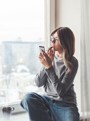 Woman with eyeglasses and curly hair paints her lips with lipstick and uses smartphone as mirror. Fast make-up. Morning routine. Cup of hot coffee on windowsill.