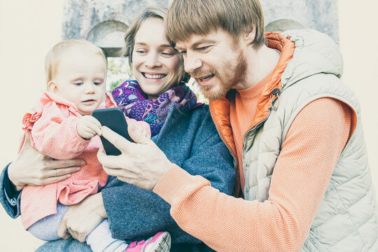 Happy parents showing picture on phone to toddler girl. Family with baby walking near old construction outdoors. Communication concept