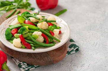 Cooked vegetables in a plate on a gray background close-up. Copy space for text. Steamed bean pods, pea pods, cauliflower, broccoli, and peppers. Healthy diet food, vegan food.