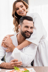 Cheerful woman in bathrobe hugging boyfriend near food on blurred foreground in hotel