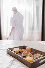 Fototapeta premium Breakfast on wooden tray on bed and couple in bathrobes embracing near window on blurred background in hotel room