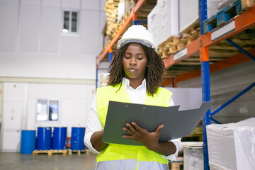 Focused Black female inspector in hardhat and safety vest walking in warehouse, carrying open...