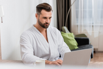 Bearded man in bathrobe using laptop near cup on blurred foreground in hotel room