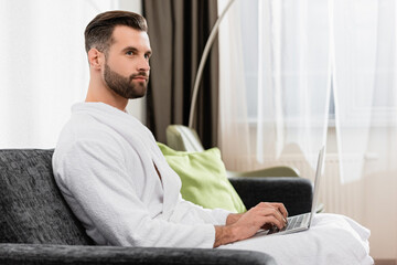 Freelancer in white bathrobe using laptop in hotel room