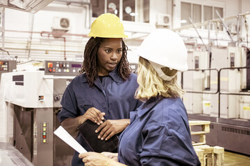 Diverse female factory employees in hardhats and overalls standing on plant floor and chatting....
