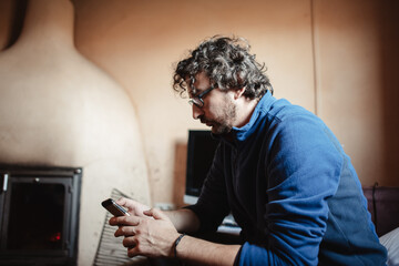 Hombre en casa de campo con chimenea mirando el teléfono móvil inteligente con sudadera azul.