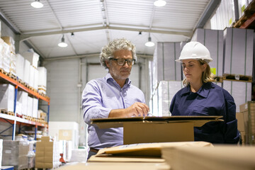 Serious safety inspector consulting female logistic employee while completing documents in warehouse. Medium shot, front view. Labor and inspection concept