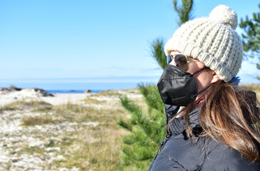 Woman with Covid-19 black face mask on a beach in Winter with black coat, sunglasses and white wool hat. KN95 or N95 or FFP2 mask.