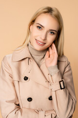 blonde woman in trench coat smiling and looking at camera isolated on beige