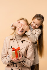 kid covering eyes of happy mother with present isolated on beige