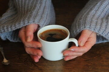 Woman's hands in sweater hold a cup of strong coffee on wooden table. Coffee fan top view background