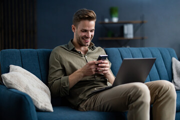 Young freelancer using smartphone and smiling. Happy man using mobile phone apps, texting message, browsing internet, looking at smartphone, sitting at home. Young people working with mobile devices.