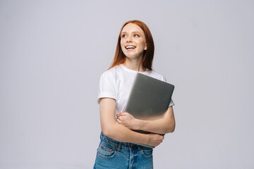 Happy young woman student holding laptop computer and looking away on isolated gray background. Pretty lady model with red hair emotionally showing facial expressions in studio, copy space.
