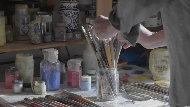 Male artist hands in a chaotic studio selecting the best paintbrush out of a pot on a sunny morning 