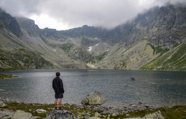 Rear view of hiking man standing and looking at lake Hincovo Pleso and foggy rocky mountains at High Tatras, Slovakia