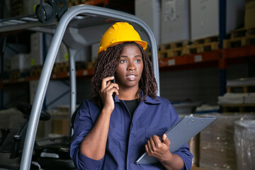 Focused Black warehouse worker in yellow hardhat standing near forklift and talking on cell....