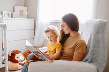 Mom reading book with baby boy toddler at home. Early age children education, development. Mother and child kid spending time together. Family authentic candid lifestyle. - obrazy, fototapety, plakaty
