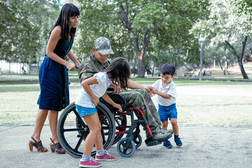 Obraz premium Disabled military father talking with cute children. Caucasian middle-aged dad in camouflage uniform outdoors with family. Pretty mom pushing wheelchair. Family reunion and veteran of war concept