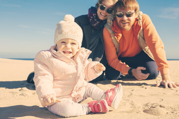 Cheerful couple watching cute baby girl wearing warm clothes, sitting on sand outdoors. Leisure time and parenthood concept