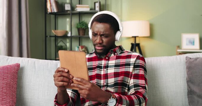 Portrait of handsome unhappy young African American man watching movie or video online in white headphones with upset face sitting on couch in cozy apartment alone using tablet device. Leisure concept
