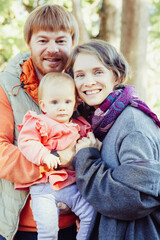 Cheerful family couple and toddler daughter posing outdoors. Parents holding cute baby girl in arms, standing in autumn park, looking at camera and smiling. Vertical shot. Family portrait concept