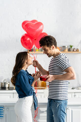 side view of happy man holding heart-shaped balloons and gift box near smiling girlfriend