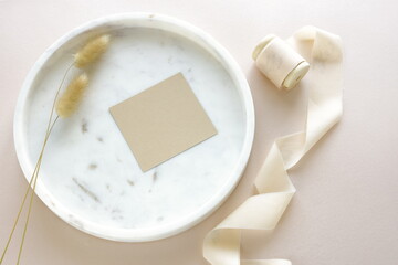blank paper sheets, greeting card mock up, ribbon, dry plant on marble tray top view with copy space. Stationery still life in beige.Flat lay.